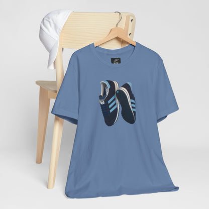 Blue t-shirt with a graphic of blue and black sneakers hanging on a wooden chair with a white cap