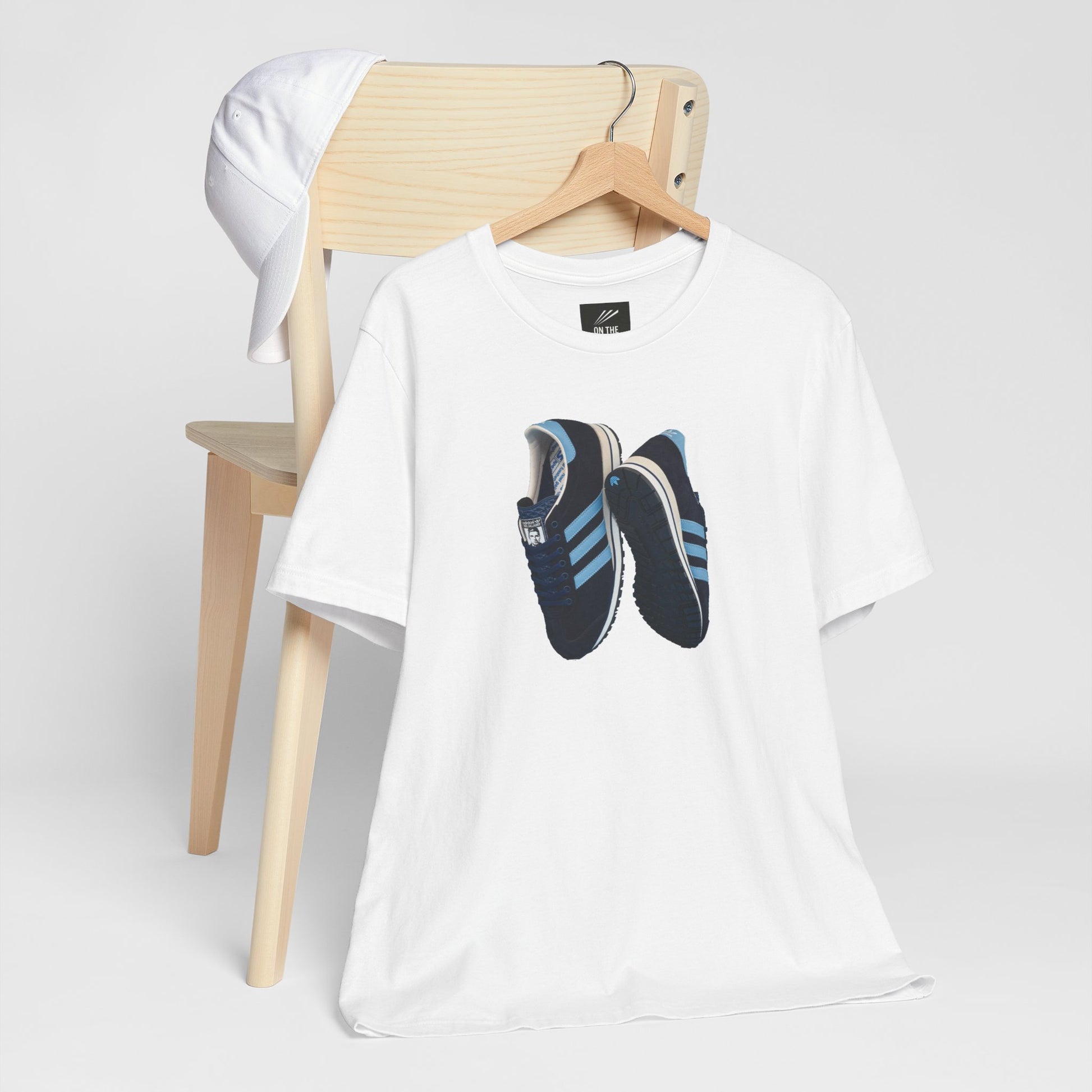 White t-shirt featuring blue and black sneakers print hanging on wooden chair with white cap