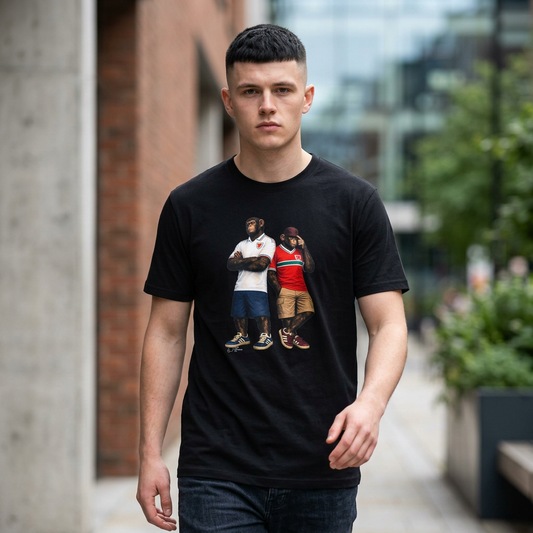 Man wearing a black short-sleeved t-shirt featuring a central graphic of two chimpanzees styled as Wales national football team supporters. The chimpanzee on the left stands with arms crossed, wearing a white away jersey with green and red trim on the collar and a red dragon crest. The chimpanzee on the right wears the classic red Wales home jersey featuring a prominent green and white horizontal chest stripe, paired with khaki cargo shorts and a burgundy bucket hat outdoors