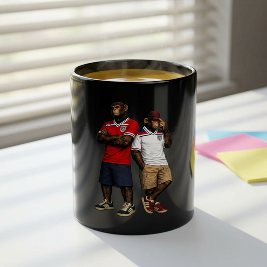 The cup shows an 11oz black ceramic mug featuring a graphic of two chimpanzees dressed in vintage England football casual attire. The monkey on the left is wearing a retro red away kit with dark blue shorts and navy trainers. The monkey on the right wears a vintage white home kit, a maroon cap, tan shorts, and red terrace-style trainers. Both characters stand against a solid black background.