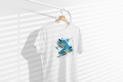 A white short-sleeve t-shirt hanging on a wooden hanger from a white rail, featuring a graphic print of blue sneakers with gold stripes and blue paint splatters on the front. The background is white with soft diagonal shadows.