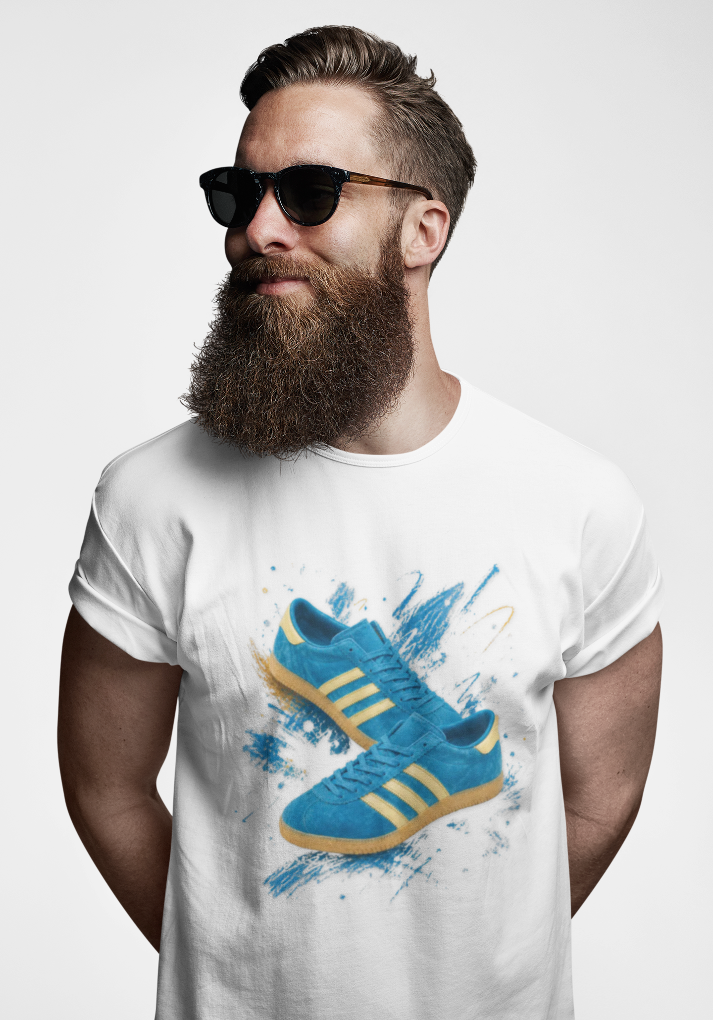 A man with a full beard and sunglasses wearing a white t-shirt with a graphic print of blue Adidas sneakers with yellow stripes, set against a plain light gray background.