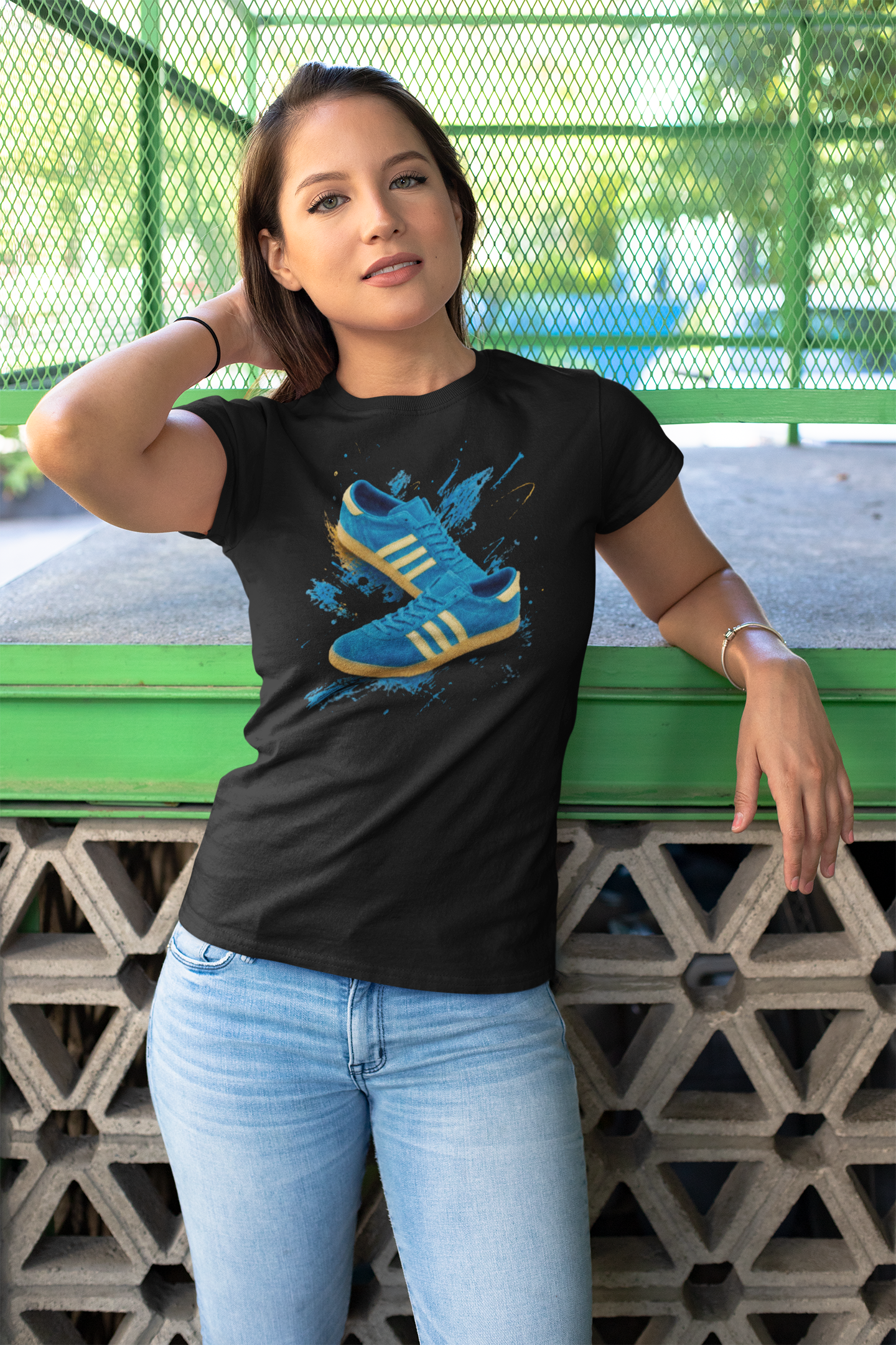 A woman wearing a black graphic t-shirt with a blue and yellow sneaker print, paired with light blue jeans, leaning against a green metal fence at an outdoor park.