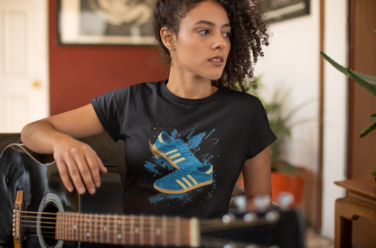 A woman with curly hair wearing a black graphic t-shirt featuring a blue and yellow sneaker design, holding an acoustic guitar in a warm indoor setting.
