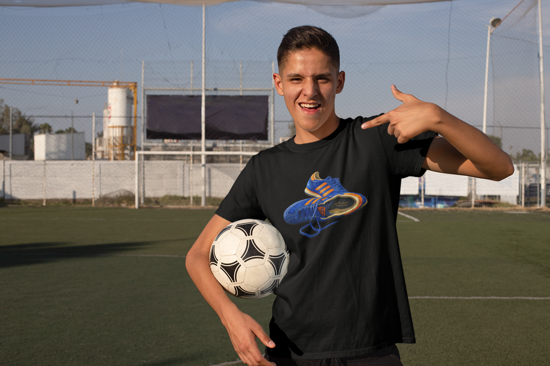 A smiling young man on an outdoor soccer field wearing a Black t-shirt with hand-illustrated blue and orange retro terrace trainer sneaker graphic print, On The Terrace casual clothing brand
