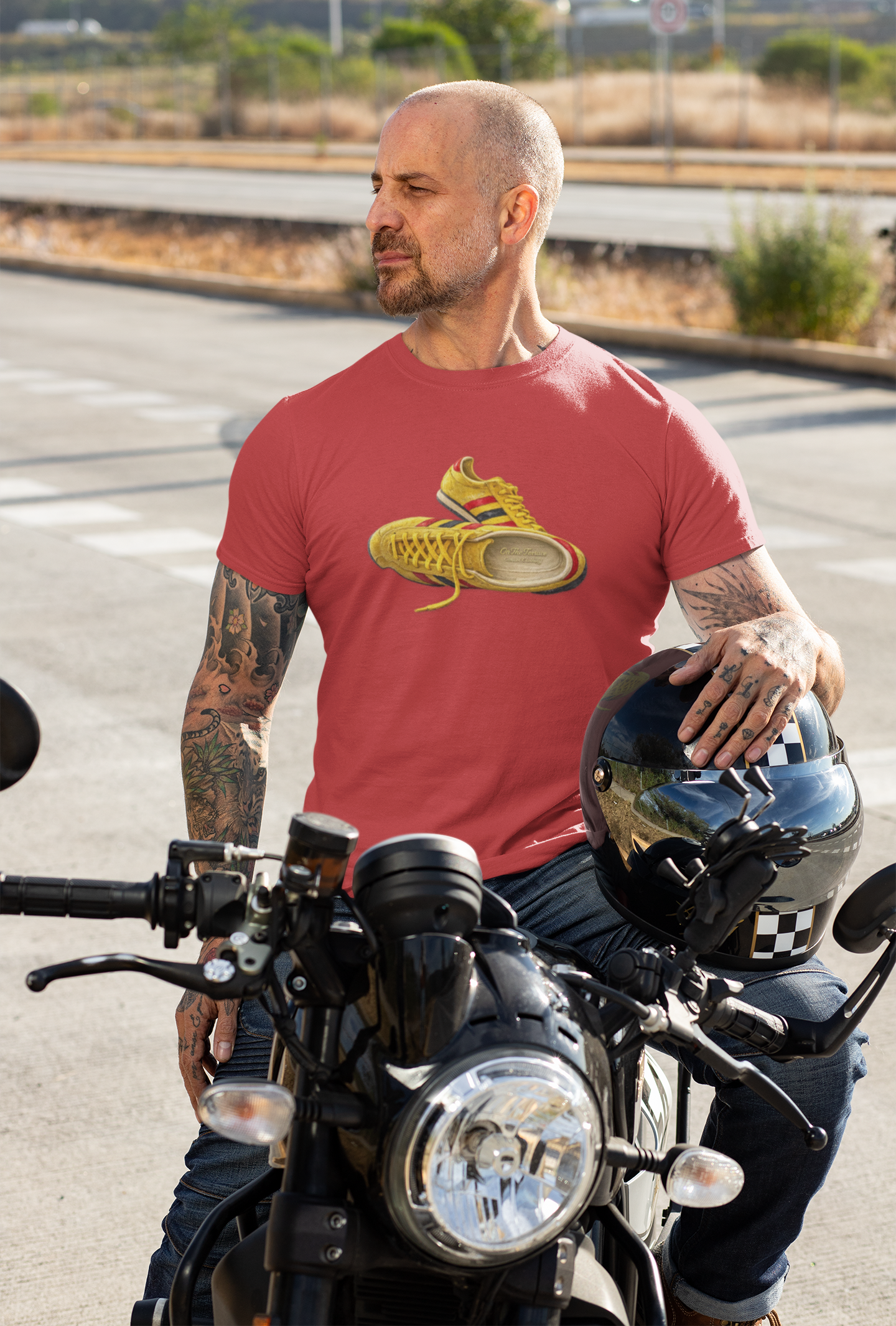 A man with tattoos sitting on a motorcycle, wearing a red t-shirt with hand-illustrated yellow, navy and red retro terrace trainer sneaker graphic print, On The Terrace casual clothing brand

