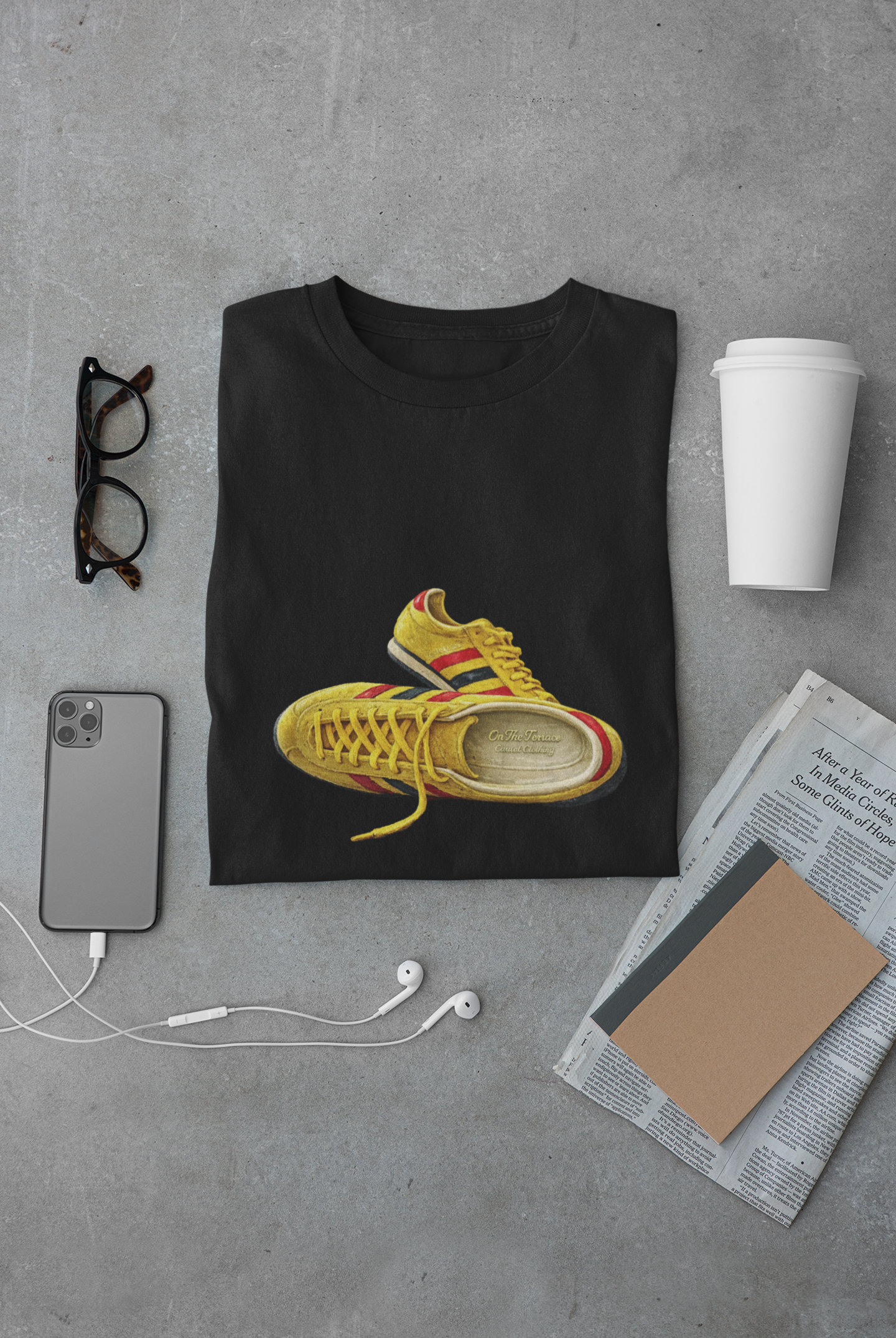 A black crewneck t-shirt with hand-illustrated yellow, navy and red retro terrace trainer sneaker graphic print, On The Terrace casual clothing brand, presented in a flat-lay arrangement on a gray concrete background with a smartphone, glasses, earbuds, a coffee cup, and a newspaper.
