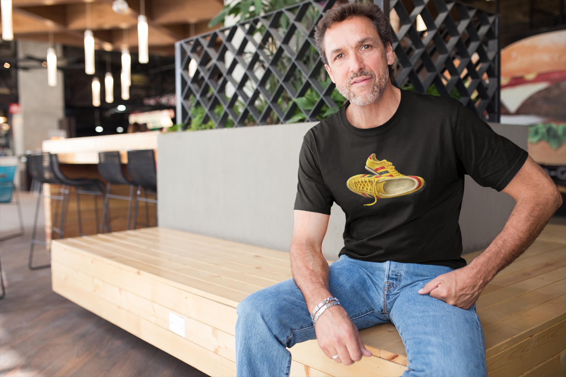 A man sitting on a wooden bench in a modern cafe setting, wearing a black t-shirt with hand-illustrated yellow, navy and red retro terrace trainer sneaker graphic print, On The Terrace casual clothing brand