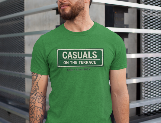 A man wearing a Hibernian green heavy cotton t-shirt flat lay featuring a central vintage green and white street sign graphic reading "CASUALS ON THE TERRACE" with "Leith • Since 1875" printed below, and an inner neck label for On The Terrace, set against a modern metal railing background.