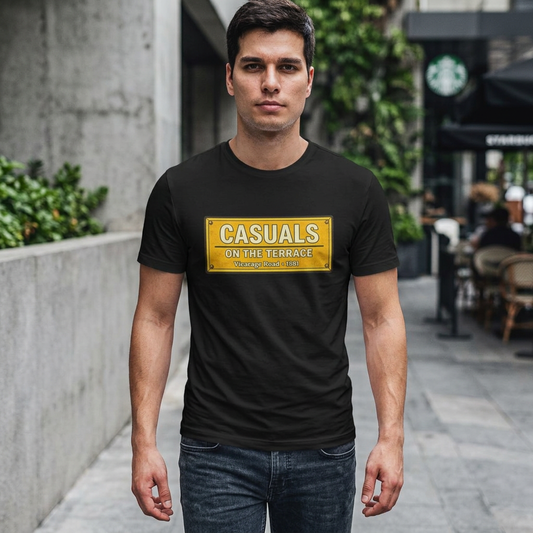 A man wearing a black Watford t-shirt with a yellow street sign graphic that reads 'CASUALS ON THE TERRACE' in a blurred outdoor urban setting.