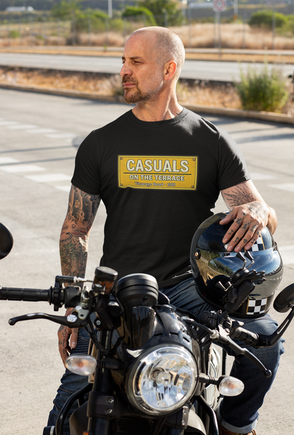 A man with tattoos sits on a motorcycle outdoors, wearing a black Watford T-shirt with a yellow rectangular patch that reads 'CASUALS ON THE TERRACE'. He is holding a black helmet with a checkered pattern.