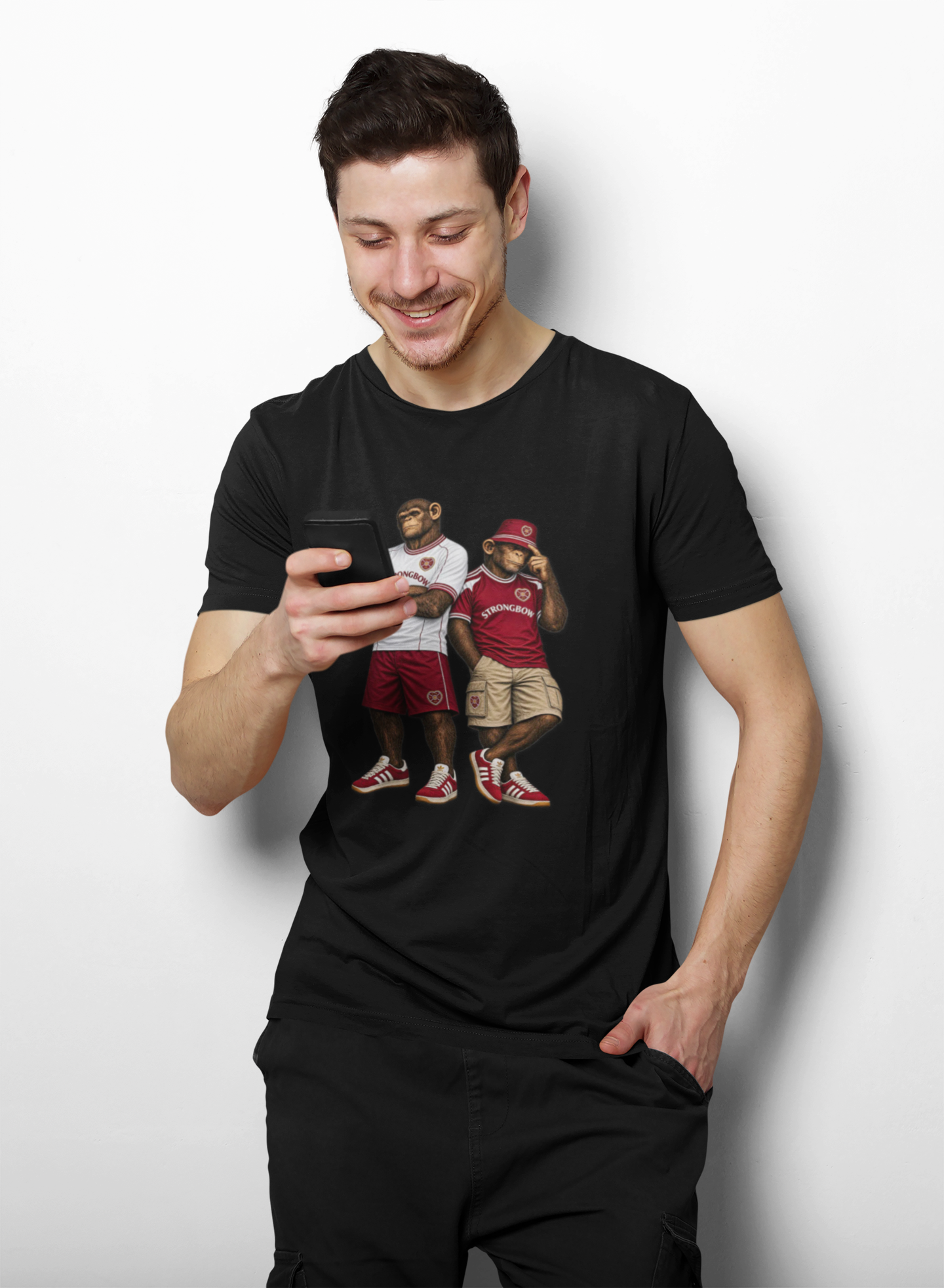 A man wearing a black Hearts t-shirt with a graphic print, looking down at a phone he is holding. The background is a clean white studio wall.