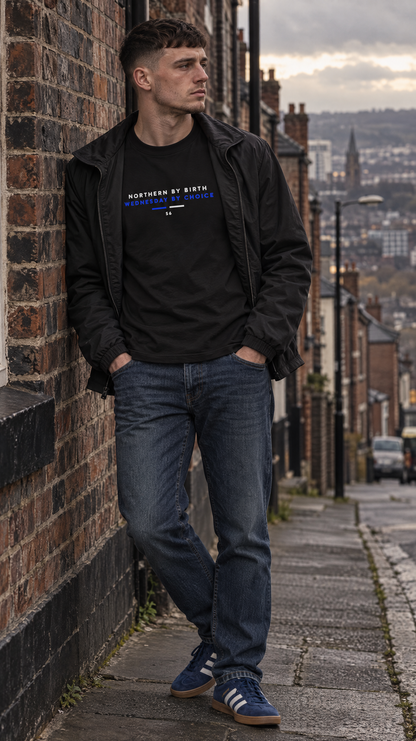 A man stands on a cobblestone street leaning against a brick wall, wearing a black graphic T-shirt under a black jacket, blue jeans, and blue sneakers. The T-shirt has white and blue text that reads 'NORTHERN BY NORTH'.