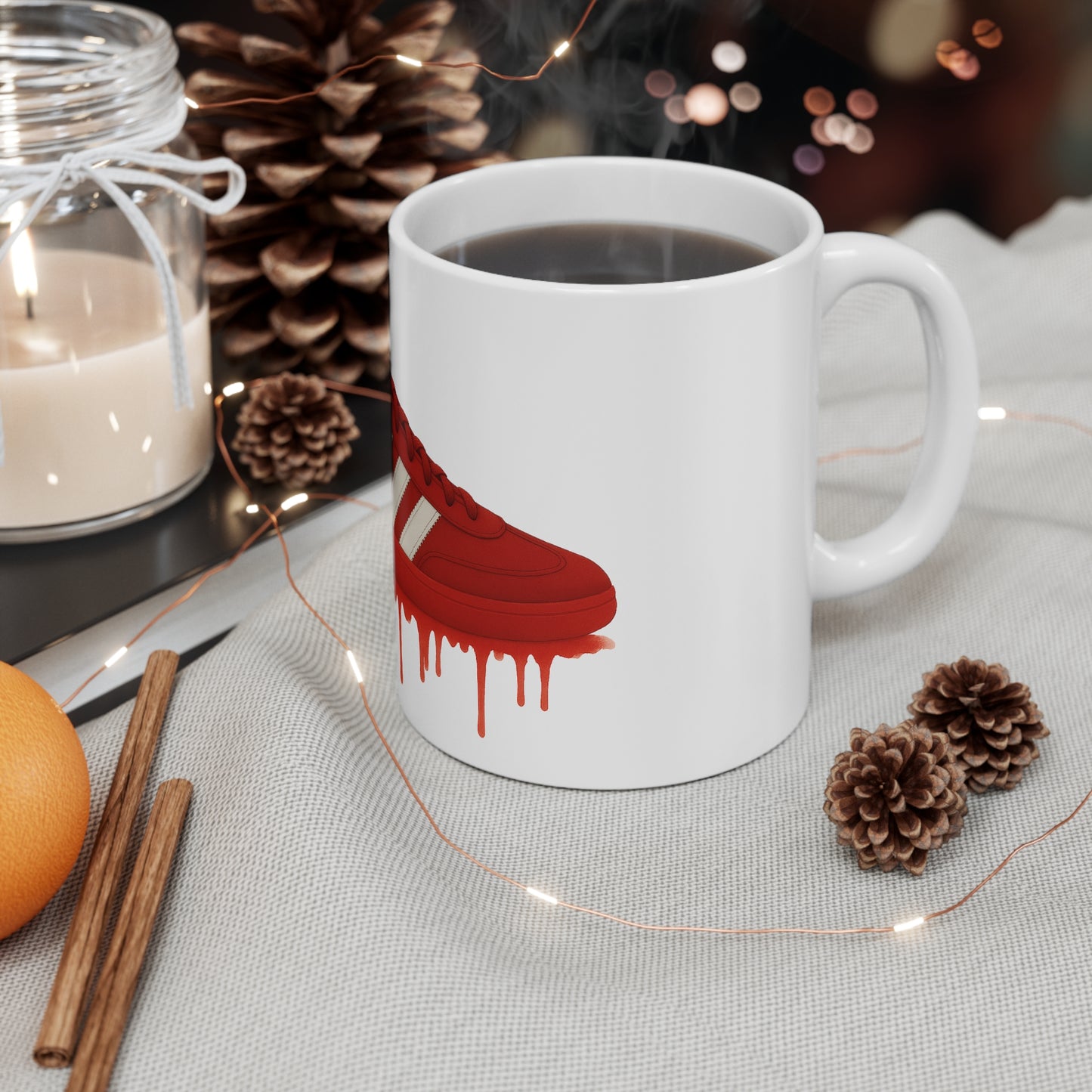 White mug with a red dripping sneaker design, filled with hot coffee, surrounded by pinecones, candle, cinnamon sticks, and orange