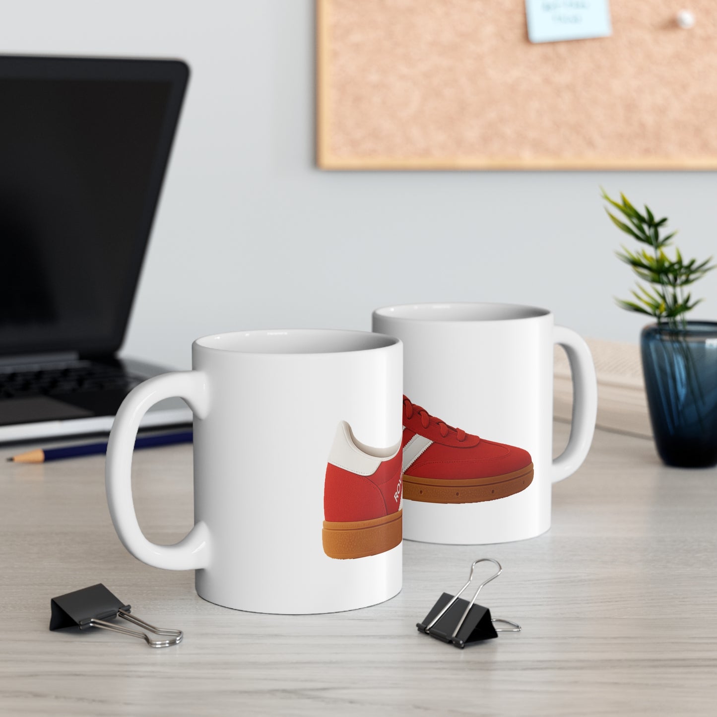 White ceramic mugs with red sneaker design on a wooden office desk with laptop, clips, and plant