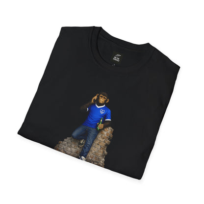 A folded black short-sleeved crew-neck t-shirt featuring a central graphic of a chimpanzee dressed in a solid blue football jersey, denim jeans, and blue trainers. The character is leaning against a section of a ruined brick wall while holding a green bottle and talking on a mobile phone.