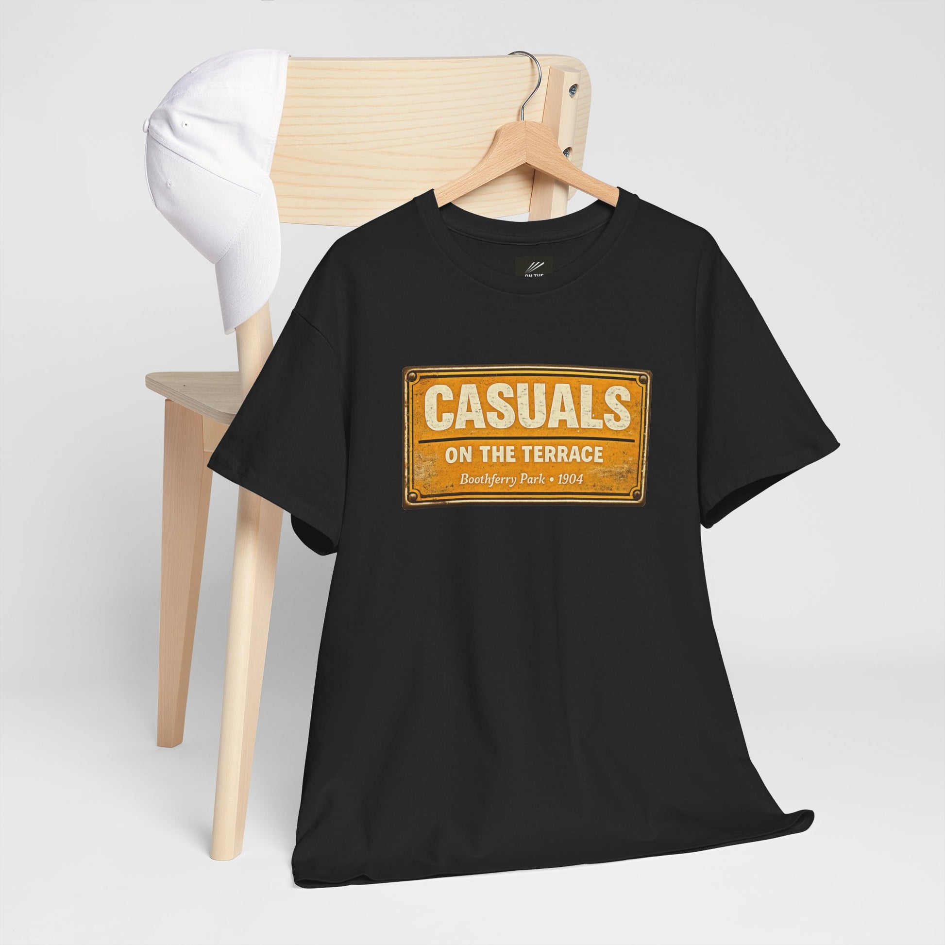 A black short-sleeve HULL CITY t-shirt on a wooden hanger, styled in front of a light wood chair with a white cap resting on it. The shirt features a vintage-style yellow and orange sign graphic on the chest that reads 'CASUALS ON THE TERRACE' with 'Bermuda Fun • 1984' underneath.