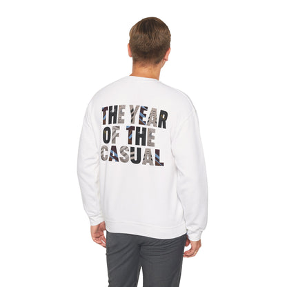 A man viewed from behind wearing a white crewneck sweatshirt with the text 'THE YEAR OF THE CASUAL' printed on the back in a black and gray patterned font, paired with dark gray pants, against a plain white background.