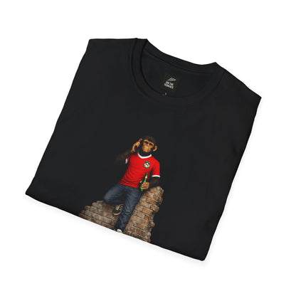 A folded black short-sleeved crew-neck t-shirt featuring a central graphic of a chimpanzee dressed in a red football jersey with white trim, denim jeans, and red trainers. The character is leaning against a section of a ruined brick wall while holding a green bottle and talking on a mobile phone.