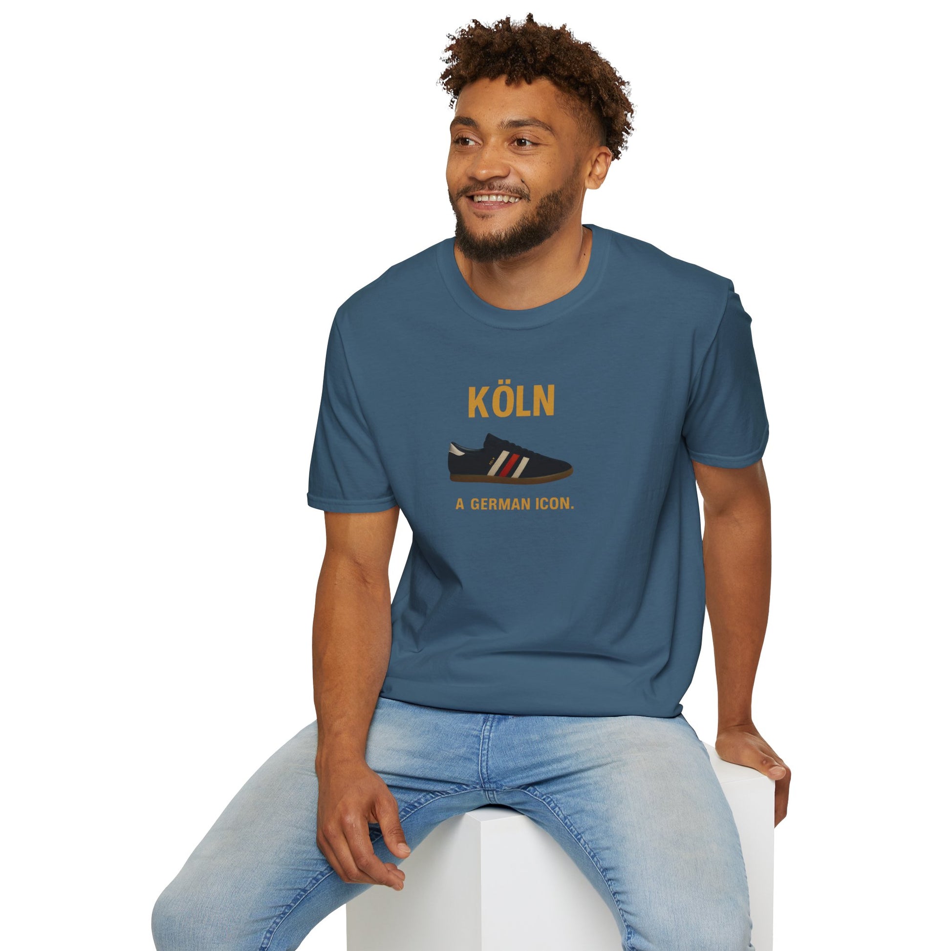 Smiling man wearing blue t-shirt with Köln German icon sneaker print, seated on white cube
