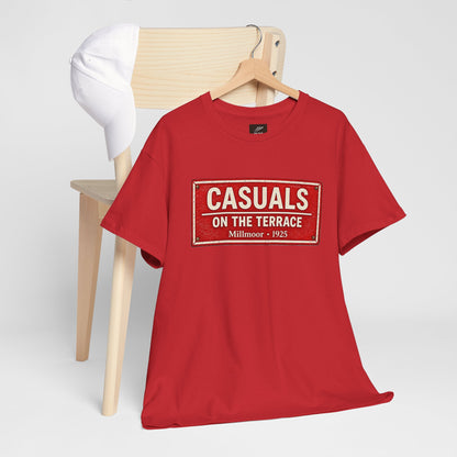 A red short-sleeve Rotherham t-shirt with a vintage-style white and red graphic reading 'CASUALS ON THE TERRACE Millmoor - 1925' hanging on a wooden hanger in front of a light wood chair with a white cap on it, set against a light gray background.