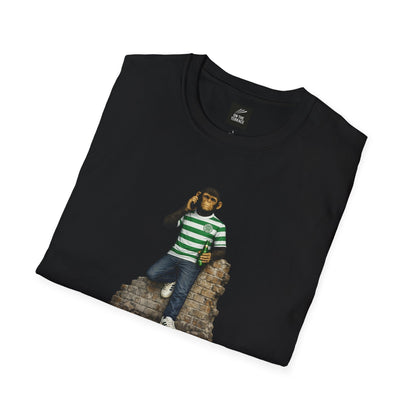 A folded black short-sleeved t-shirt featuring a central graphic of a chimpanzee wearing a green and white hooped football jersey, denim jeans, and white trainers. He is leaning against a section of a ruined brick wall while holding a green bottle and talking on a mobile phone.