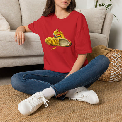 A woman sitting cross-legged on a woven rug in front of a light beige sofa, wearing a red t-shirt with hand-illustrated yellow, navy and red retro terrace trainer sneaker graphic print, On The Terrace casual clothing brand
