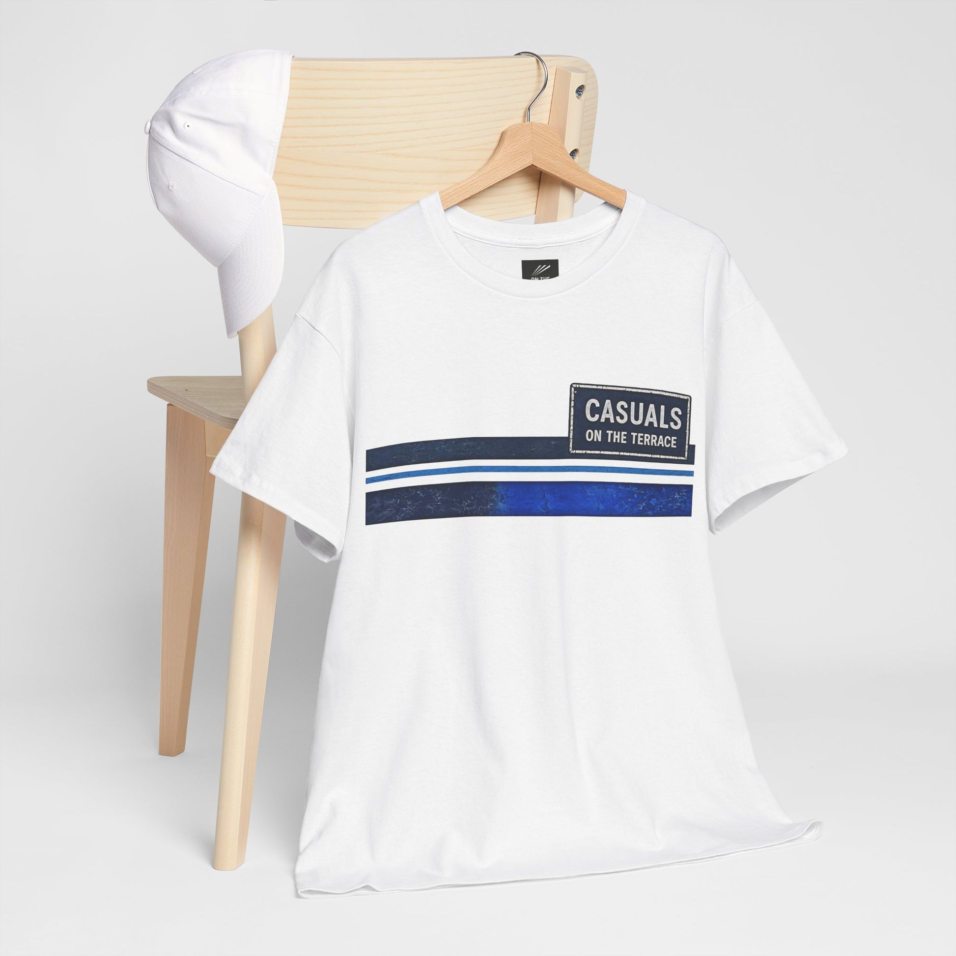 A white short-sleeve t-shirt with a graphic stripe design on the chest, hanging on a wooden hanger from a light wood chair. A matching white baseball cap is resting on the back of the chair.