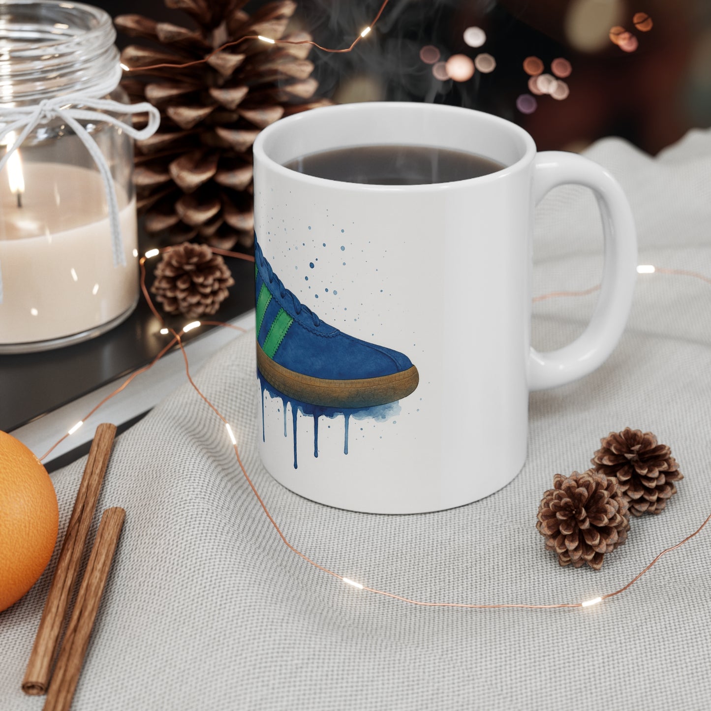 Steaming coffee mug with blue sneaker art on a cozy cloth surrounded by pine cones, candle, cinnamon sticks