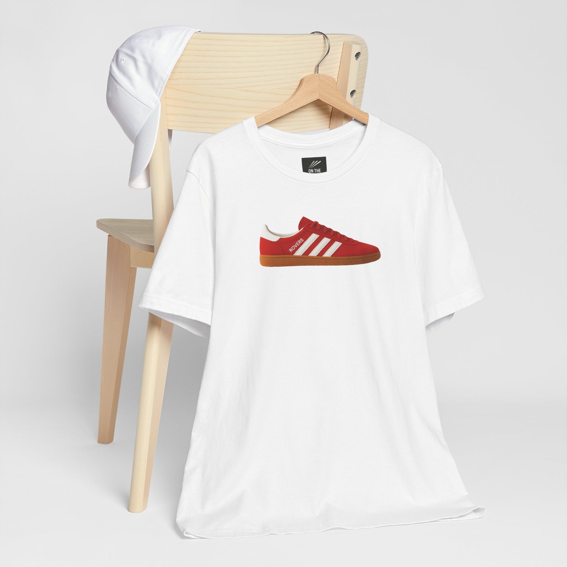 White t-shirt with red sneaker graphic and white cap on wooden chair on white background