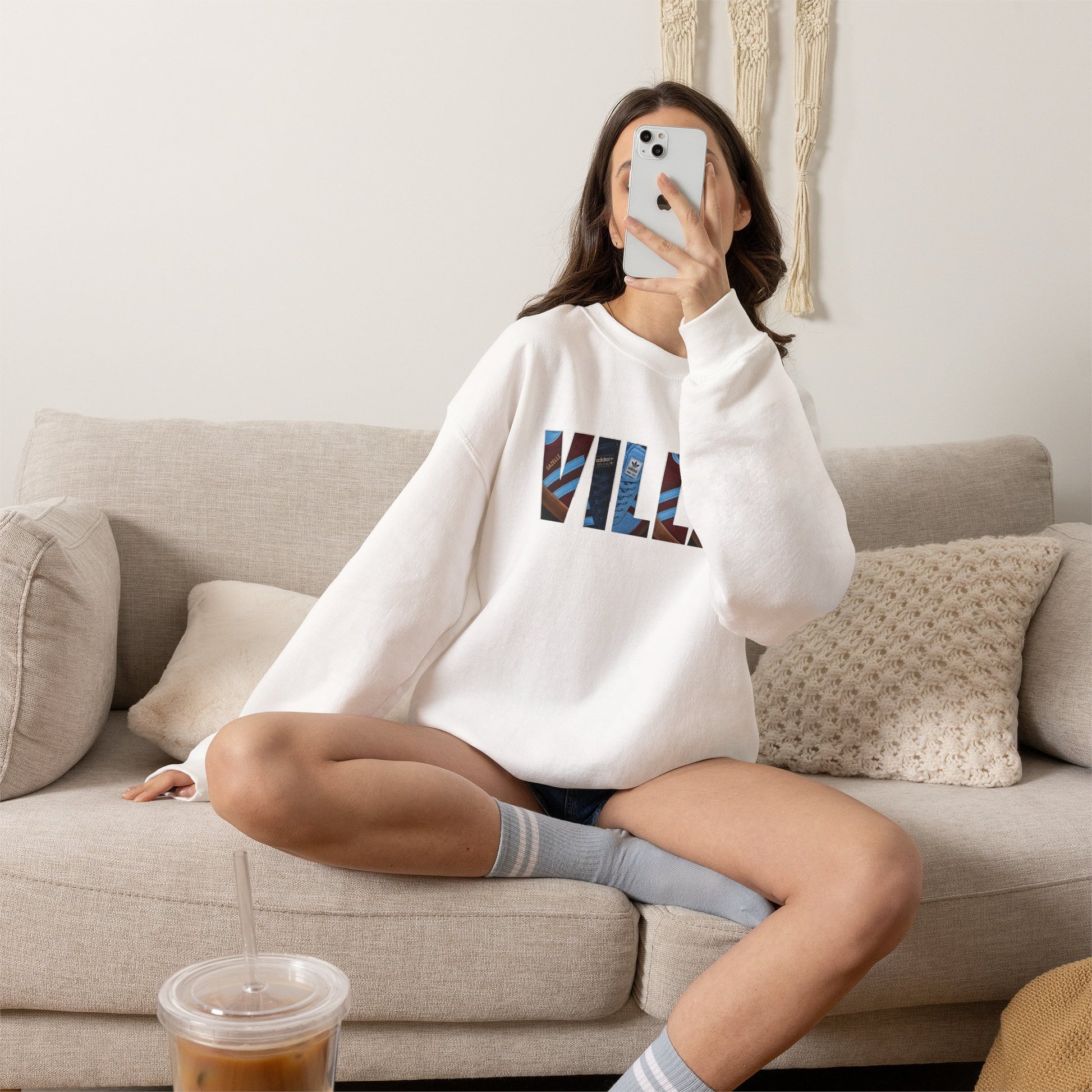 A woman sits cross-legged on a beige couch taking a mirror selfie while wearing a white sweatshirt with VILLA lettering.
