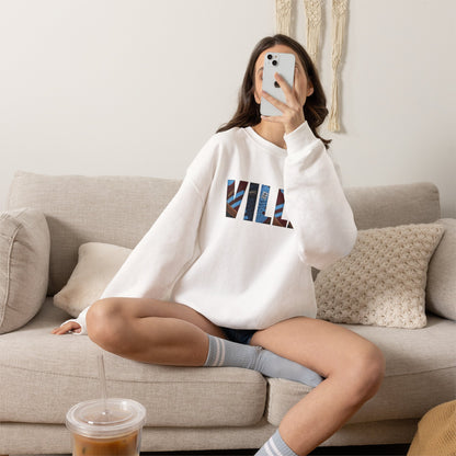 A woman sits cross-legged on a beige couch taking a mirror selfie while wearing a white sweatshirt with VILLA lettering.