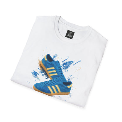 White t-shirt with a graphic print of a blue suede trainer with cream three stripes and gum sole, set against a blue and gold paint-splash background, On The Terrace neck label visible.