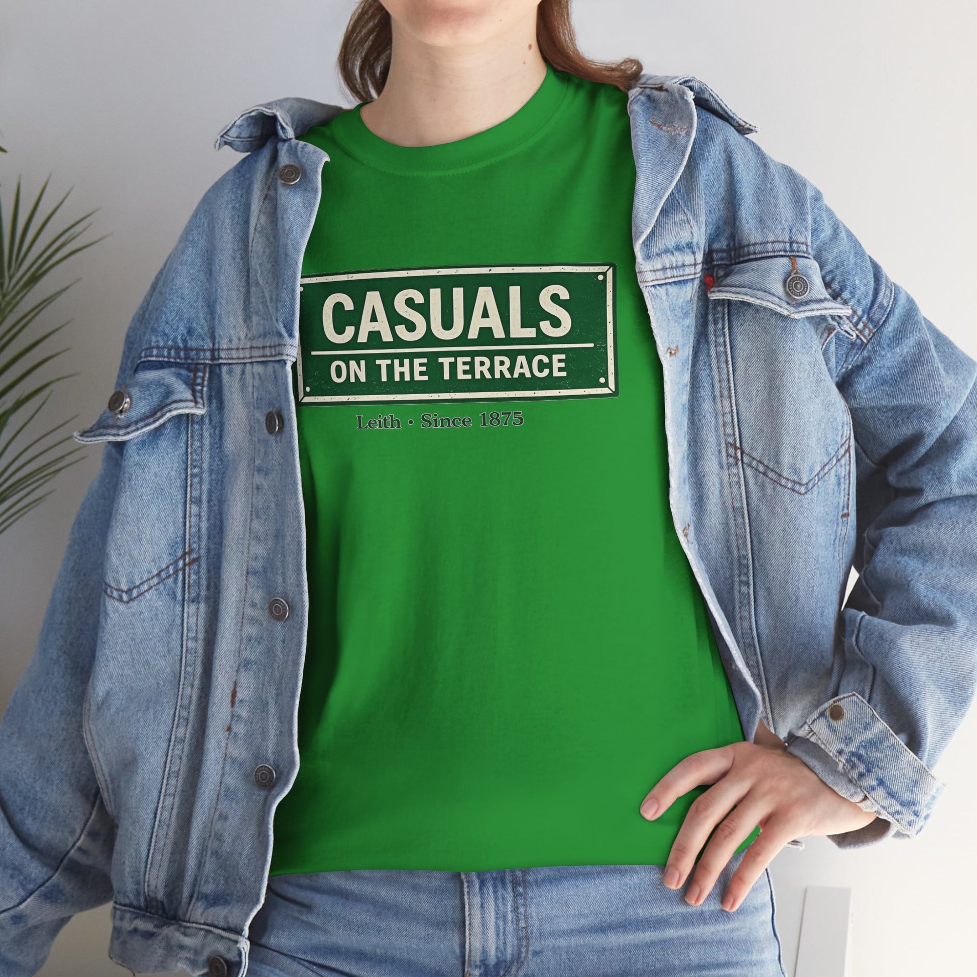 A person wearing a Hibernian green heavy cotton t-shirt flat lay featuring a central vintage green and white street sign graphic reading "CASUALS ON THE TERRACE" with "Leith • Since 1875" printed below, and an inner neck label for On The Terrace in a street-sign style print.