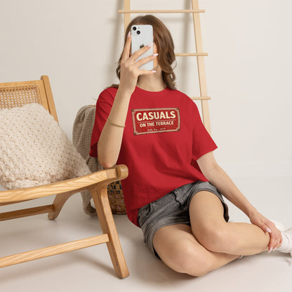 Person wearing a red DRFC t-shirt flat lay featuring a central vintage red and white street sign graphic reading "CASUALS ON THE TERRACE" with "Belle Vue • 1879" printed below, and an inner neck label for On The Terrace., sitting on the floor with a wooden chair and ladder in the background.