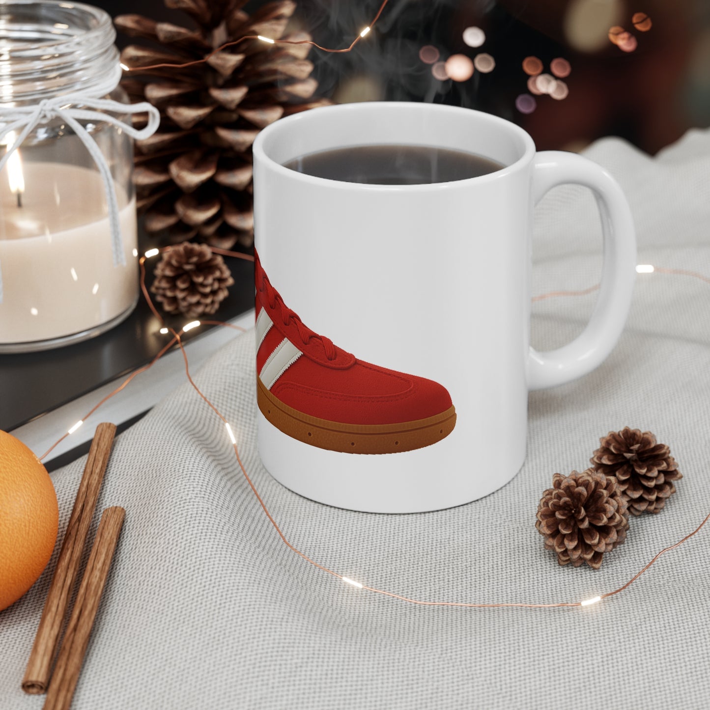 White coffee mug with red sneaker design on cozy table with pine cones, candle, cinnamon sticks, and orange