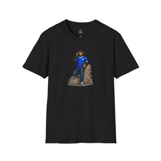 A black short-sleeved crew-neck t-shirt featuring a central graphic of a chimpanzee dressed in a solid blue football jersey, denim jeans, and blue trainers. The character is leaning against a section of a ruined brick wall while holding a green bottle and talking on a mobile phone.