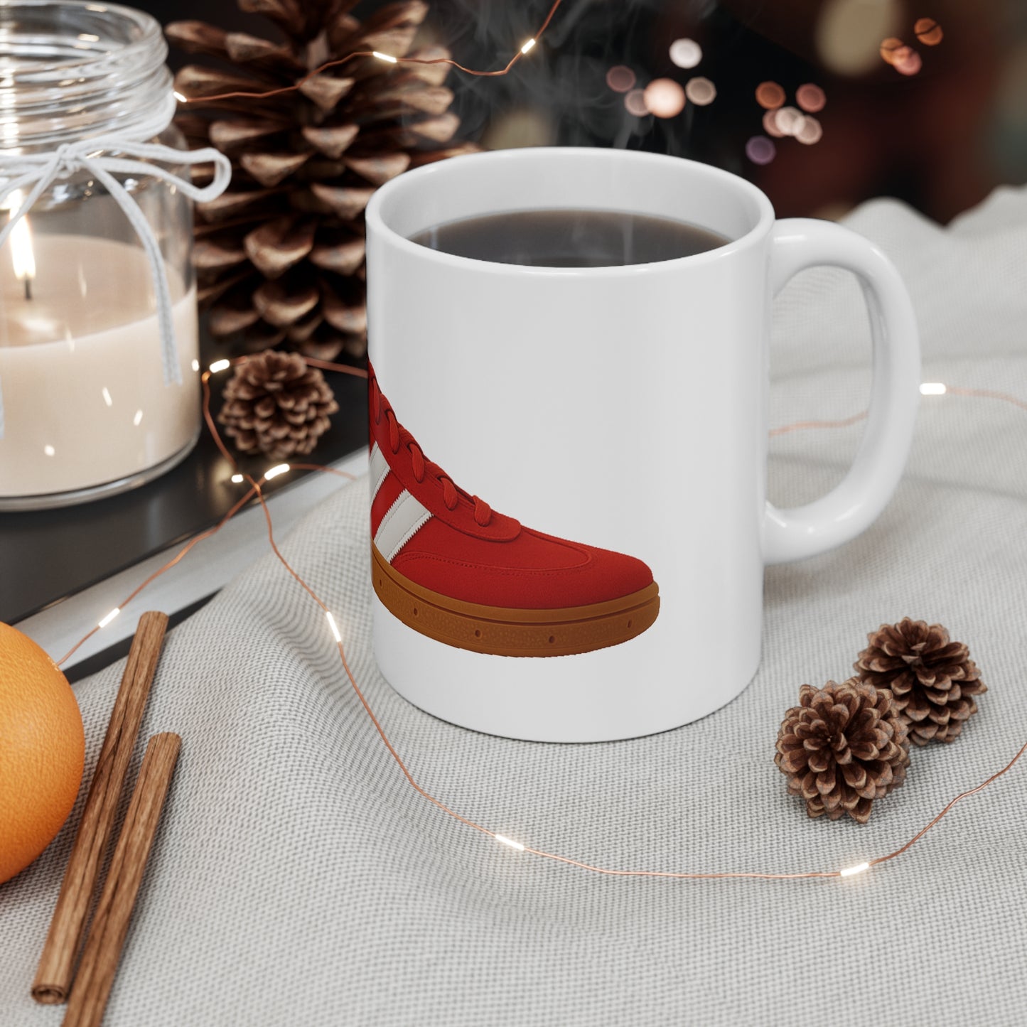 White mug with red sneaker design filled with steaming coffee, surrounded by pinecones, cinnamon sticks, orange, and candle