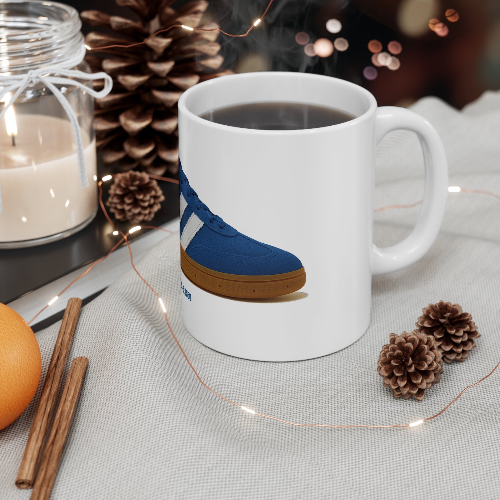 White coffee mug with a blue sneaker illustration, surrounded by pine cones, a lit candle, cinnamon sticks, and orange on a fabric surface