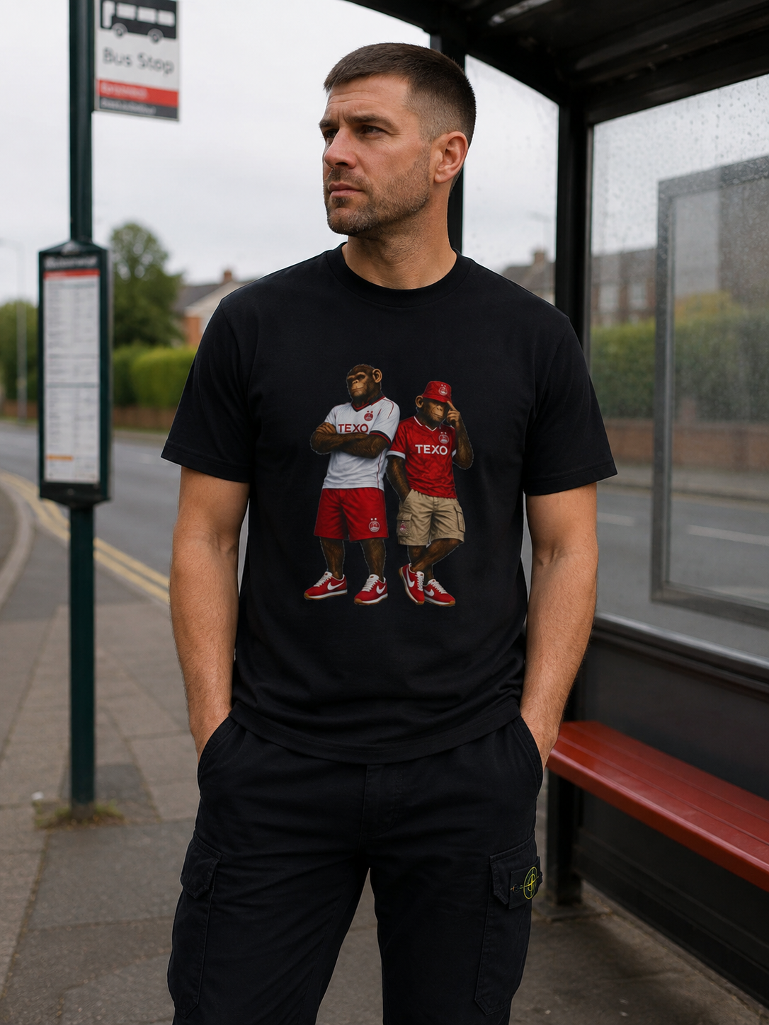 A man standing at a bus stop wearing a black crewneck Aberdeen t-shirt with a graphic print of two bears dressed in Aberdeen kits