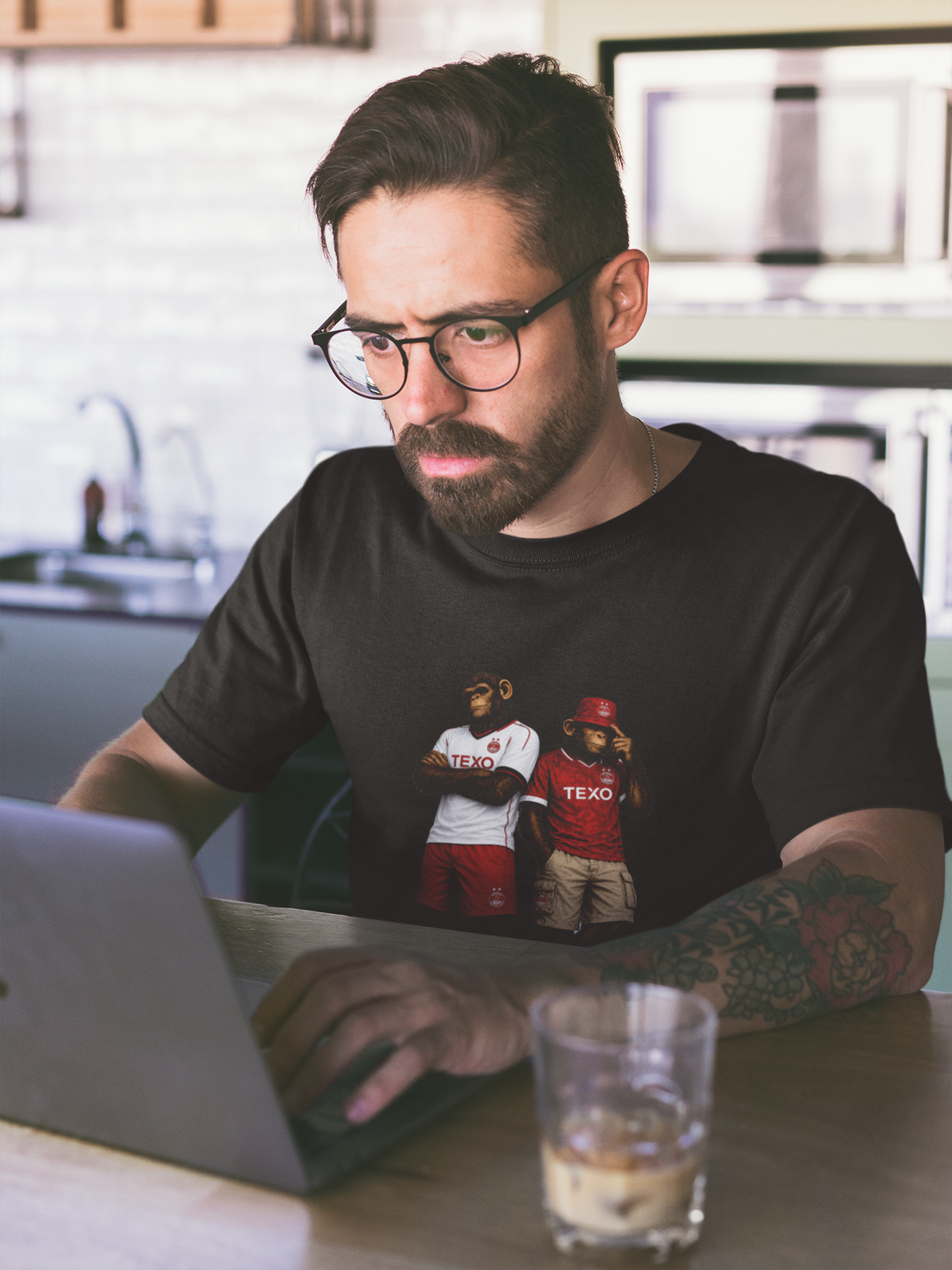 A man with glasses and a beard sits at a table in a kitchen setting, working on a laptop. He is wearing a black Aberdeen t-shirt with a graphic print of two cartoon characters wearing TEXO jerseys.