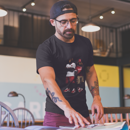 A man wearing a black graphic Aberdeen t-shirt with a printed design of two men, standing in a cafe-like environment and looking down at papers on a table.