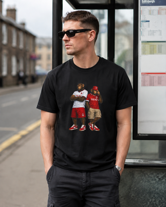 A man wearing a black graphic Aberdeen t-shirt with a humorous print of two Aberdeen players in red and white jerseys, standing outdoors near a bus stop.