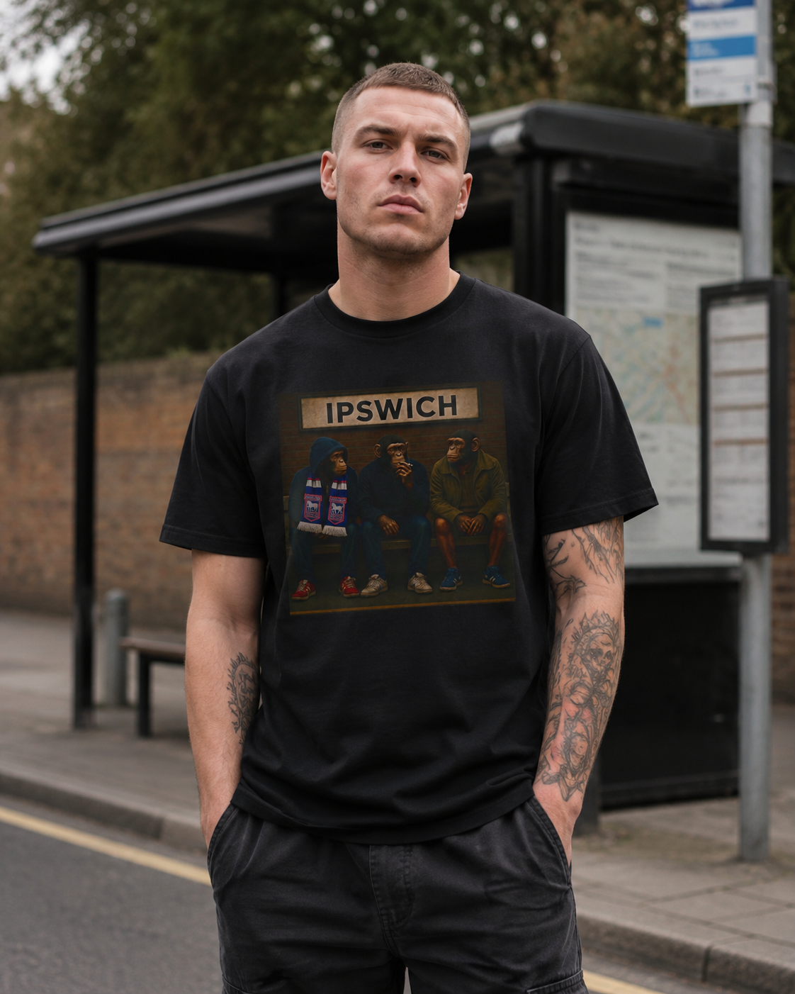 A man standing at a bus stop wearing a black Ipswich t-shirt with a vintage-style print of the band IPSWICH and dark cargo pants.