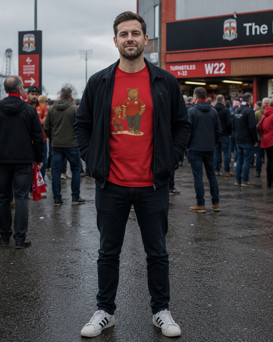 A man standing outdoors in front of a stadium or club building, wearing a red Barnsley graphic T-shirt with a teddy bear design, a black zip jacket, dark jeans, and white sneakers.