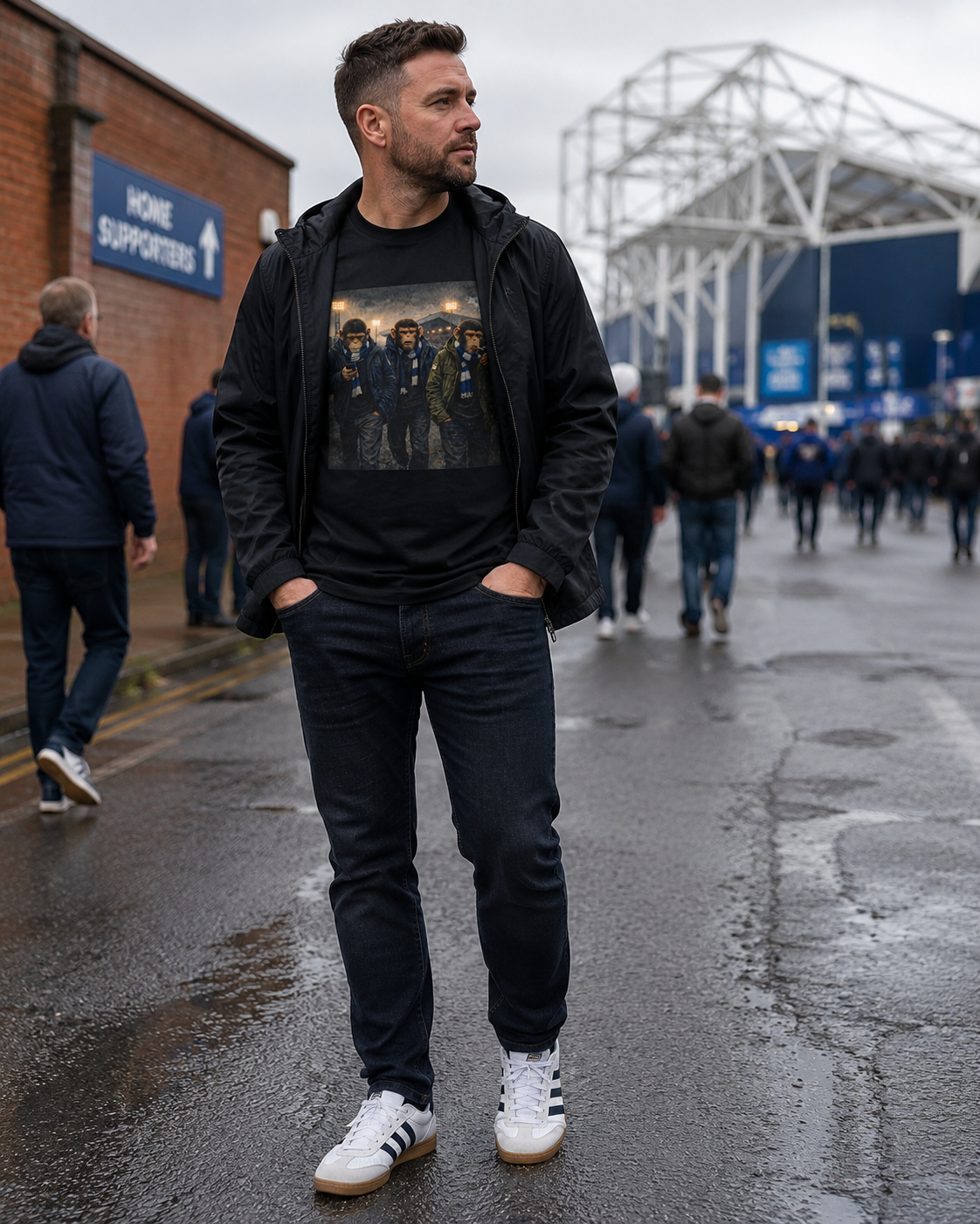 A man stands on a wet street outside a stadium wearing a black graphic t-shirt under a black zip-up hoodie, dark jeans, and white sneakers. The background shows the stadium and other people walking.