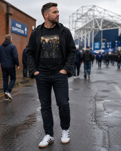 A man stands on a wet street outside a stadium wearing a black graphic t-shirt under a black zip-up hoodie, dark jeans, and white sneakers. The background shows the stadium and other people walking.