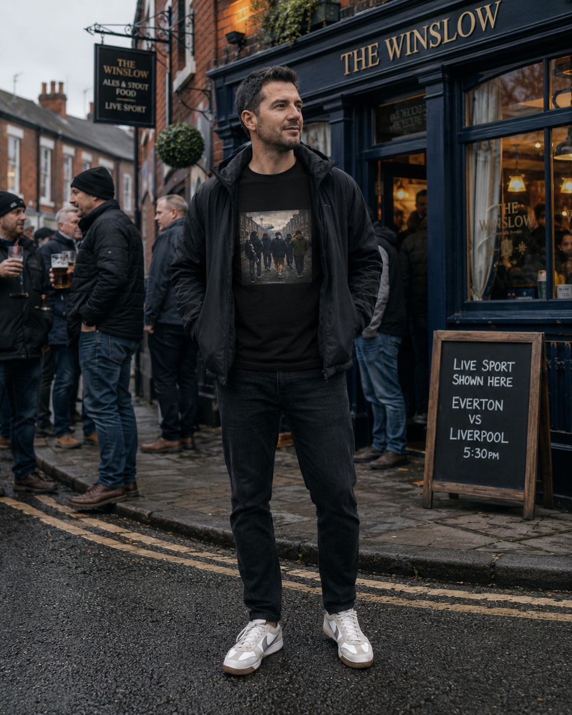 A man wearing a black Northern Monkey graphic t-shirt under a black zip-up hoodie and black jeans stands on the street in front of 'The Winslow' pub. He is wearing white sneakers.