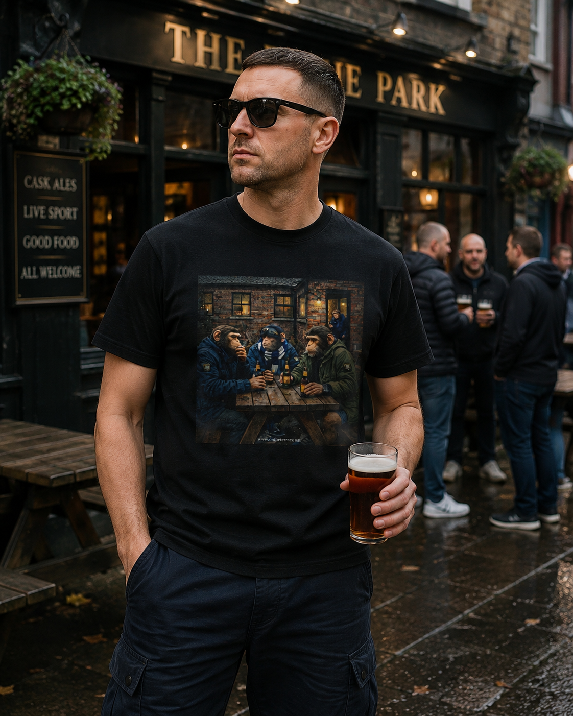 A man wearing a black Northern Monkey graphic t-shirt with a detailed print of a pub scene, standing outside 'The Park' pub and holding a pint of beer.
