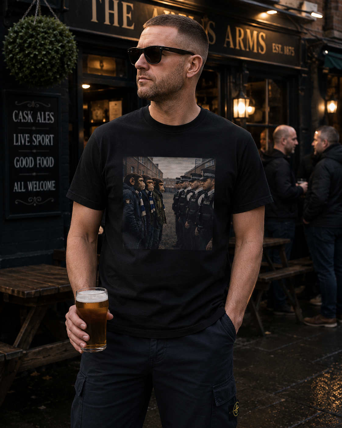 A man wearing a black graphic t-shirt and dark cargo pants stands outside a pub holding a pint of beer. The t-shirt features a rectangular graphic print of soldiers in formation.